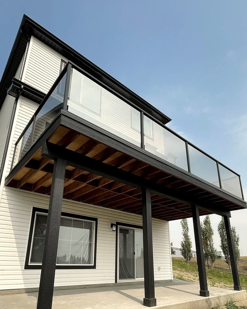 A raised deck with a glass railing and black wrapped posts over a concrete patio at the back of a two storey house