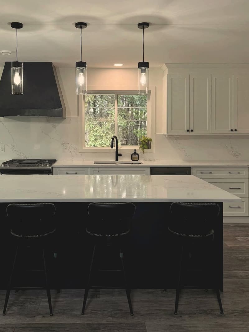 A renovated kitchen with white cabinets, a black island with a quartz top and three glass pendant lights over the window