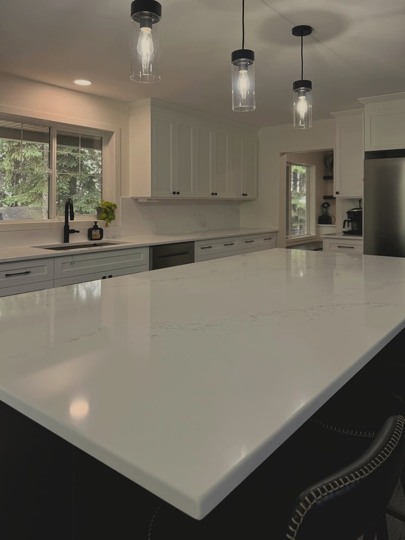 A white quartz island top under three glass pendant lights, with white cabinets and a window over the sink